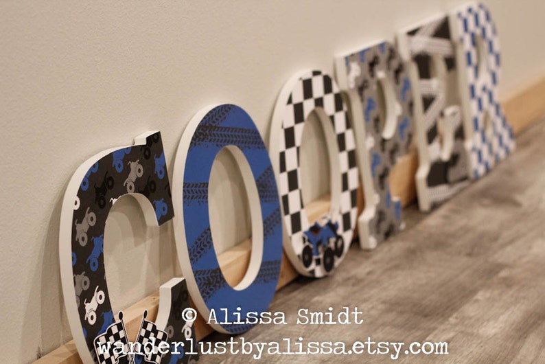 May include: Wooden letters spelling the name "COOPER" decorated with black and white checkered, blue and black tire tread, and gray and black monster truck patterns.