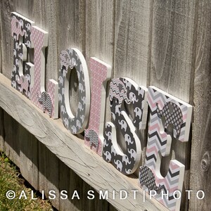 May include: Wooden letters spelling out the name "Eloise" in pink and gray. Each letter is decorated with a different pattern of pink and gray elephants.