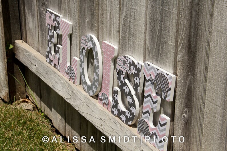 May include: Wooden letters spelling out the name "Eloise" in pink and gray. Each letter is decorated with a different pattern of pink and gray elephants.