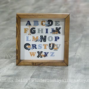 May include: A framed alphabet chart with a white background and a brown wooden frame. The letters are in blue and yellow with construction vehicles and tools surrounding each letter.