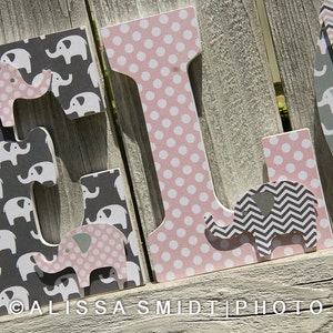 May include: Three wooden letters spelling "ELI" decorated with pink and gray elephant patterns. The letter "E" has a pink elephant with white polka dots, the letter "L" has a pink background with white polka dots, and the letter "I" has a gray elephant with a chevron pattern.