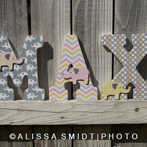 May include: Wooden letters spelling the name "MAX" in gray, pink, and yellow. Each letter is decorated with a different pattern of elephants, polka dots, and chevron stripes.