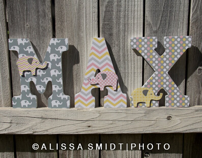 May include: Wooden letters spelling the name "MAX" in gray, pink, and yellow. Each letter is decorated with a different pattern of elephants, polka dots, and chevron stripes.