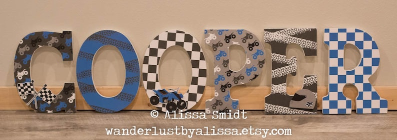 May include: Wooden letters spelling the name "COOPER" decorated with a racing theme. The letters are painted in blue, black, white, and gray with checkered patterns, tire tracks, and small race car graphics.