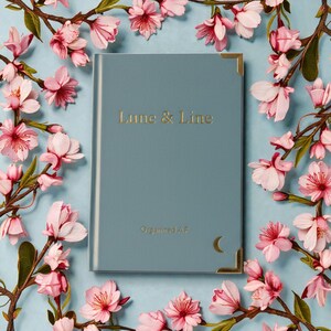 May include: A blue journal with gold lettering that reads "Lune & Line" and "Organized AF". The journal has gold corner protectors and a crescent moon symbol. Pink and white flowers surround the journal on a light blue background.