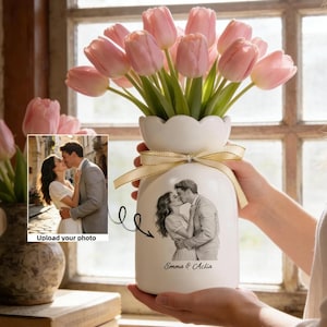 May include: A white ceramic vase with a scalloped edge, holding pink tulips and a beige ribbon. The vase features a black and white sketch of a kissing couple, with the names "Emma & Aclia" below. A photo with the text "Upload your photo" is also visible.