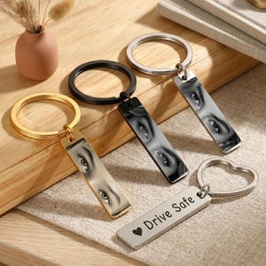 May include: Four metal keychains with rectangular tags. Three tags feature black and white close-up prints of human eyes. One silver-tone tag is engraved with the text Drive Safe and a heart symbol. The keychains have gold, black, and silver rings.