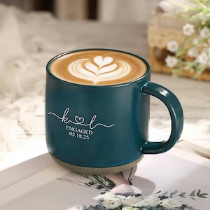 Può includere: Una tazza in ceramica verde acqua con un latte macchiato con un disegno a forma di cuore. La tazza ha un manico e una base grigia. La scritta bianca sulla tazza recita "k♡l ENGAGED 05.18.25".