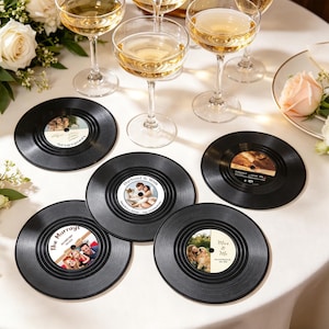 May include: A collection of black vinyl record coasters with personalized photo inserts. Champagne glasses and floral arrangements surround the coasters on a white tablecloth. The coasters feature wedding and family photos.