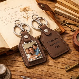 May include: Two brown leather keychains with silver metal hardware. One keychain features a rectangular photo insert showing a man, a girl, and a boy smiling. The other keychain is engraved with the text, I need you, I love you, Dad, in a dark font.