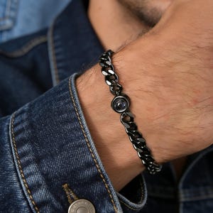 Può includere: Un bracciale a catena gourmette in metallo nero con un ciondolo circolare dal centro scuro e riflettente. Il bracciale è composto da maglie lucide intrecciate che formano una fascia continua, caratterizzata da una finitura scura ed elegante.