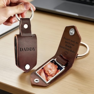May include: A brown leather keychain with the word "DADDY" engraved. The keychain has a silver ring and a photo of a baby. The second keychain has the text "I can't wait to meet you Daddy Love from Baby" and a heart symbol.