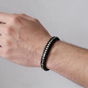 Könnte beinhalten: Schwarzes Lederarmband mit silberfarbenen Perlen und Metallstäben. Das Armband wird am Handgelenk getragen und hat ein geflochtenes Design. Die Perlen und Metallstäbe sind gleichmäßig verteilt. Ein stilvolles Accessoire.