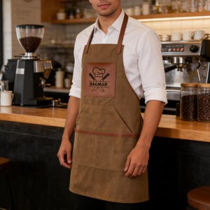 May include: Brown apron with leather straps and a front pocket. The apron features a leather patch with a chef's hat, crossed knives, and the name "DAGMAR". The apron is worn by a person in a white shirt.