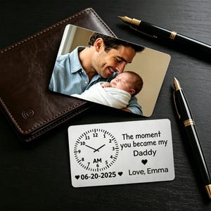Könnte beinhalten: Eine personalisierte Metall-Geldbörsenkarte mit einem Foto von Vater und Baby und einem Zifferblatt-Design. Die Karte lautet "Der Moment, in dem du mein Papa wurdest" mit Datum und Namen. Eine braune Lederbrieftasche und zwei Stifte sind ebenfalls sichtbar.