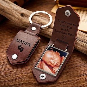 Könnte beinhalten: Ein brauner Lederschlüsselanhänger mit Metallring, der außen das Wort DADDY und ein Fußabdrucksymbol zeigt. Die Innenklappe ist mit dem Text I can't wait to meet you Daddy Love from Baby und einem Herz graviert und enthält ein Baby-Ultraschallbild.
