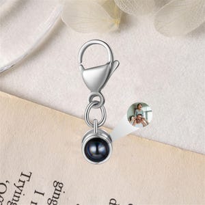 May include: A silver-colored charm with a lobster clasp, two connecting rings, and a round pendant. The pendant has a dark, reflective surface. A small inset image shows a family.