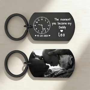 Op de afbeelding: Twee zwarte sleutelhangers. Eén sleutelhanger heeft een klokontwerp met de tekst "The moment you became my Daddy Leo" en de datum "11-24-2025". De andere sleutelhanger heeft een zwart-wit foto.