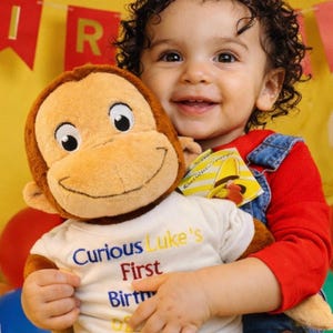 Puede incluir: Un niño sonriente con una camisa roja de manga larga y un peto vaquero sostiene un peluche de Curious George. El peluche lleva una camiseta blanca que dice "Curious Luke's First Birthday". Una pancarta amarilla con letras rojas dice "HAPPY BIRTHDAY" en el fondo.