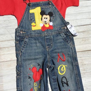 May include: A blue denim overall outfit with a red short-sleeved shirt. The overalls have a yellow number one with a Mickey Mouse applique. The overalls also have the name "Noah" embroidered on the leg in red and yellow thread.