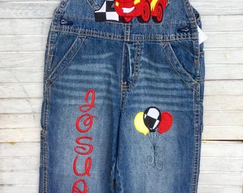 May include: Blue denim overalls with a red race car and the number 2 applique. The word "Jose" is embroidered in red on the leg, and the cuffs have a black and white checkered pattern. Three balloons are also embroidered on the leg.
