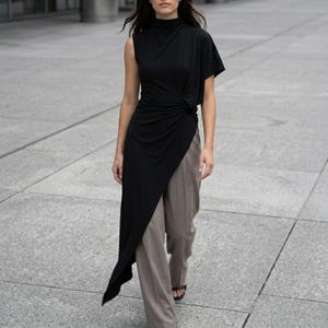May include: A black asymmetrical top with a high neckline and a draped design. The top is paired with taupe-colored trousers. The outfit is worn by a person walking on a gray concrete surface.