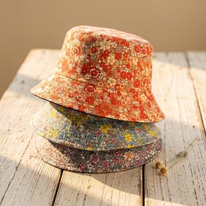 May include: A stack of three bucket hats featuring floral patterns. The top hat is orange and red, the middle hat is blue and yellow, and the bottom hat is purple and pink. Each hat has a downward-sloping brim and a rounded crown design.