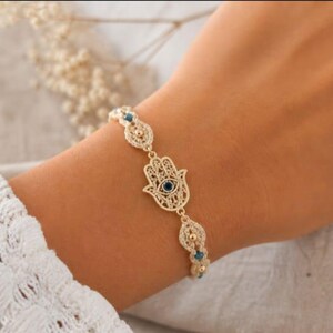 May include: Gold-toned bracelet featuring a Hamsa hand charm with a blue center stone. The bracelet has intricate detailing and is adorned with additional blue accents. The jewelry is displayed on a wrist, showcasing the design and craftsmanship.