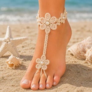 Puede incluir: Una sandalia descalza de ganchillo color crema con un diseño floral y detalles de perlas. La sandalia se lleva en un pie en una playa de arena, con conchas marinas y una estrella de mar en el fondo. El diseño incluye una flor en el tobillo y otra en los dedos.