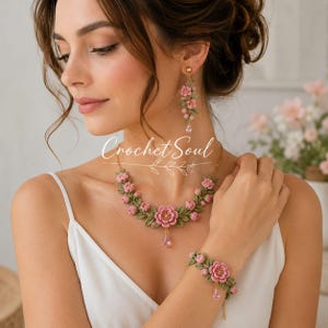 Könnte beinhalten: Ein Schmuckset bestehend aus einer Halskette, einem Armband und einem einzelnen Ohrring, gefertigt aus gehäkelten rosa Blumen und grünen Blättern. Halskette und Armband haben kleine rosa tropfenförmige Anhänger. Der Text Crochet Soul ist zu sehen.