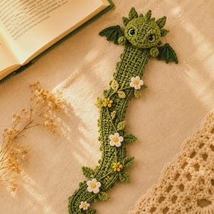Può includere: Un segnalibro a forma di drago lavorato all'uncinetto in verde, con una testa dettagliata dagli occhi scuri, piccole ali e un corpo lungo decorato con fiori all'uncinetto bianchi e gialli e foglie verdi lungo la struttura in filato.