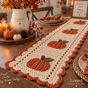 Puede incluir: Un camino de mesa rectangular de ganchillo en color crema con un patrón repetitivo de calabazas naranjas con tallos y hojas verdes. El camino tiene un borde festoneado naranja y un diseño de punto calado decorativo en los bordes.