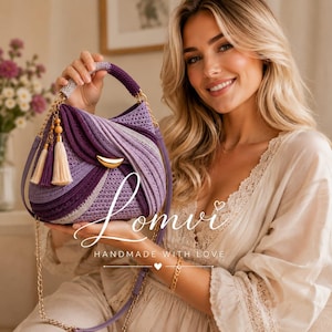 Puede incluir: Un bolso de ganchillo en tonos morados y lavanda con un detalle dorado en forma de media luna, un asa superior, una correa de cadena dorada extraíble y borlas decorativas con cuentas de madera. El bolso muestra el texto Lomvi HANDMADE WITH LOVE.