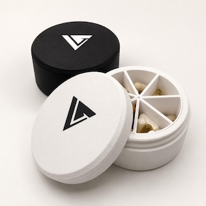 Könnte beinhalten: Zwei runde Pillendosen, eine schwarze und eine weiße, jeweils mit einem geometrischen Logo auf dem Deckel. Der weiße Behälter ist geöffnet und zeigt sechs Innenfächer mit verschiedenen beigen und weißen Kapseln zur täglichen Aufbewahrung.