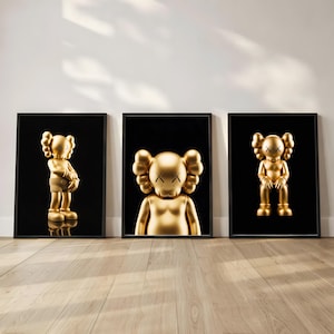 May include: Three framed art prints featuring golden sculptures against a black background. The sculptures have distinctive features, including crossed-out eyes and rounded ears. The prints are displayed on a wooden floor against a white wall.