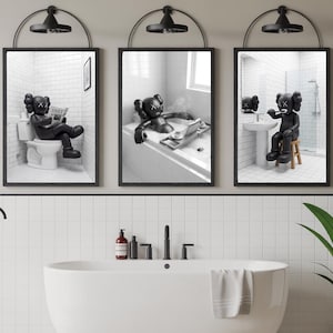 May include: Three framed black and white prints depicting a cartoon figure in bathroom scenes. One shows the figure on a toilet reading, another in a bathtub with a laptop, and the third brushing its teeth. Each print is under a black metal light fitting.