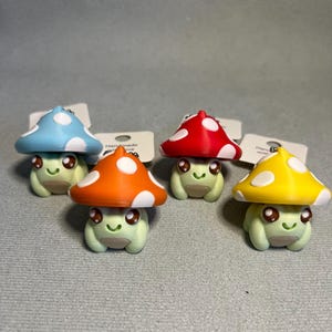 Può includere: Quattro portachiavi a forma di fungo in blu, rosso, arancione e giallo. Ogni portachiavi presenta un corpo verde chiaro a forma di rana con un volto sorridente e un cappello a fungo con pois bianchi. Fatto a mano.