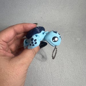 Op de afbeelding: Een lichtblauw en donkerblauw gearticuleerd schildpadfiguurtje met een gladde afwerking, voorzien van grote zwarte ogen met witte accenten en donkerblauwe stippen op de flippers. Een klein zilverkleurig metalen kogeltjesketting zit aan de kop.