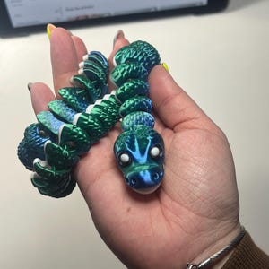 Op de afbeelding: Een gearticuleerd slangenspeelgoed in groen- en blauwtinten, vastgehouden in een hand. De kop van de slang heeft witte ogen en een blauw-groene gradiënt. De segmenten zijn gedetailleerd met een textuurpatroon. Het speelgoed is een leuk, kleurrijk item.