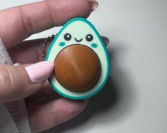 Puede incluir: Un llavero en forma de aguacate verde claro y turquesa con un hueso marrón. El aguacate tiene una cara sonriente con ojos negros y una boca pequeña. El llavero está sujeto a una cadena de bolas plateada.