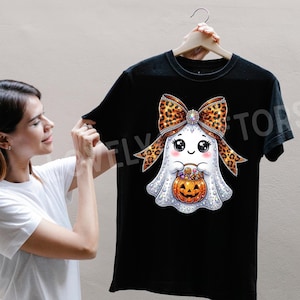Può includere: Una t-shirt nera con la grafica di un fantasma bianco con grandi occhi e guance rosate. Il fantasma indossa un grande fiocco leopardato e tiene in mano un secchiello a forma di zucca arancione pieno di caramelle colorate, in stile scintillante.