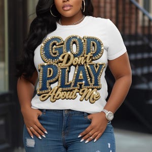 May include: A white short-sleeved t-shirt featuring the text "GOD Don't PLAY About Me" in a large, three-dimensional style. The letters have a dark blue denim-textured fill and are outlined with sparkling gold-toned rhinestones.