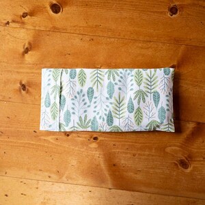 Puede incluir: Un artículo rectangular de tela con un fondo blanco y un patrón repetido de hojas y ramas verdes y turquesas. El artículo está doblado y parece una pequeña almohada o envoltura, descansando sobre una superficie de madera.