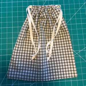 May include: A drawstring bag made of green and white checkered fabric. The bag has a gathered top with white drawstrings. The bag is on a green cutting mat.