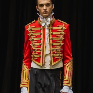 Op de afbeelding: Een rode jas in militaire stijl met gouden details en versieringen. De jas heeft gouden versieringen, een hoge kraag en een witte vlinderdas. Het model draagt een wit overhemd en een zwarte broek, wat een formele uitstraling creëert.