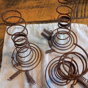 May include: Close-up of several rusty metal springs, likely from an antique bed frame. The springs are arranged on a white cloth, with a wooden table in the background. The springs have a spiral design.