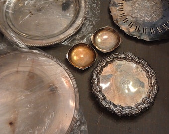 May include: A collection of silver-plated serving dishes and bowls. The dishes have a variety of decorative patterns and are arranged on a gray surface.