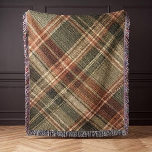 May include: A rectangular woven throw blanket featuring a diagonal plaid pattern in shades of olive green, dark brown, and cream. The blanket has a textured appearance with frayed, multi-colored fringe edges along the top and bottom sides.