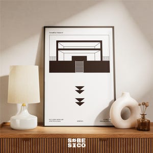 May include: A modern interior design print in a black frame, featuring a minimalist architectural illustration in black and white. A white lamp with a beige shade and a white vase with a circular opening are on a wooden surface.
