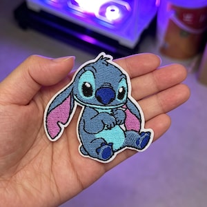 May include: An embroidered patch featuring the character Stitch in shades of blue and light blue with pink inner ears, a dark blue nose, and black eyes. The patch has a white border and depicts the character in a seated position with arms folded.